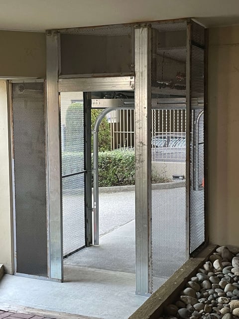 Metal-framed security door entrance with mesh screening, opening to an outdoor patio area with white railings and green foliage