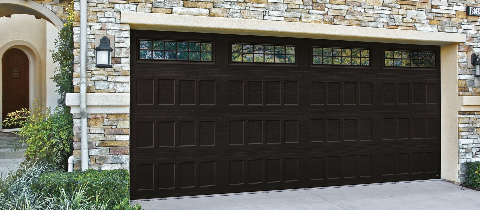 Black two-car garage door with windows on a stone and beige house facade with landscaping