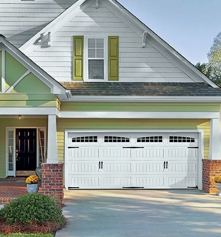 Modern two-story house with white garage doors, green shutters, brick foundation, and manicured landscaping