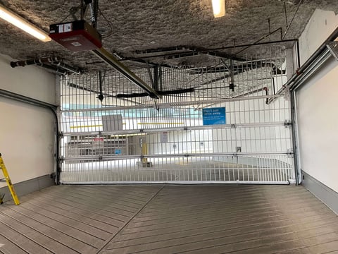 Empty garage interior with white overhead door, gray flooring, exposed ceiling, and wall-mounted storage shelves