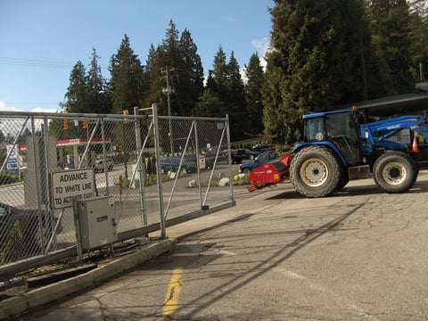 Blue tractor with red attachment approaching open metal gate at facility entrance with evergreen trees in background