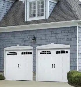 Two white garage doors with black panel windows on a gray shingled house with a peaked roof