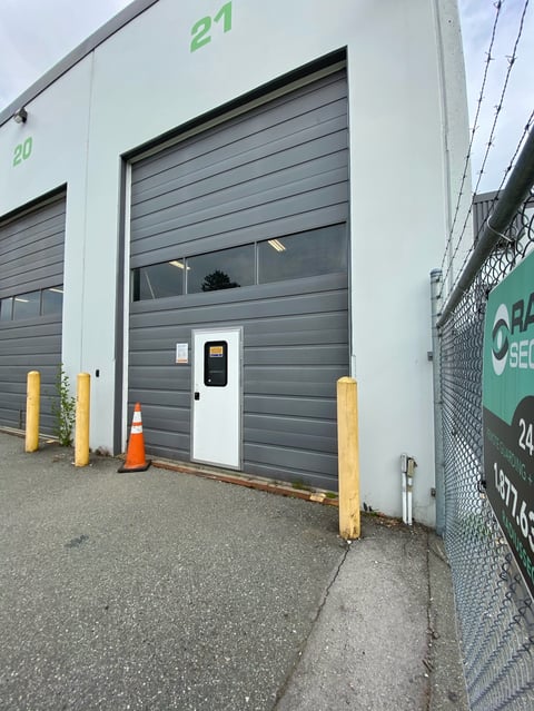 Industrial garage door with white pedestrian entrance door, numbered 27, flanked by yellow protective posts and orange traffic cone