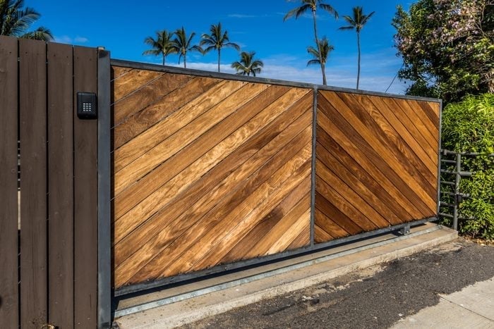 Modern wooden driveway gate with diagonal slat design and dark frame, set against palm trees and blue sky