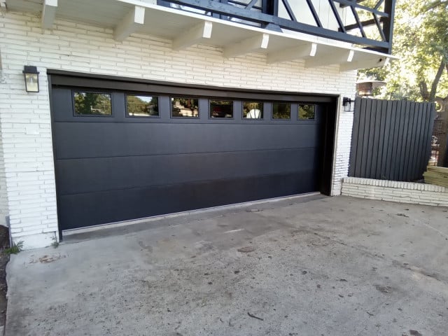 Modern black garage door with multiple windows on white brick building with concrete driveway