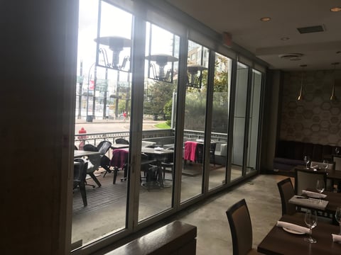 Interior view of a building with large glass doors and windows overlooking an outdoor patio seating area with tables and chairs