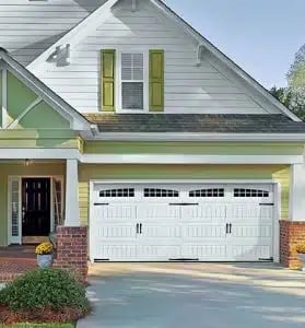 Two-story house with white garage doors, green shutters, brick foundation, and landscaping