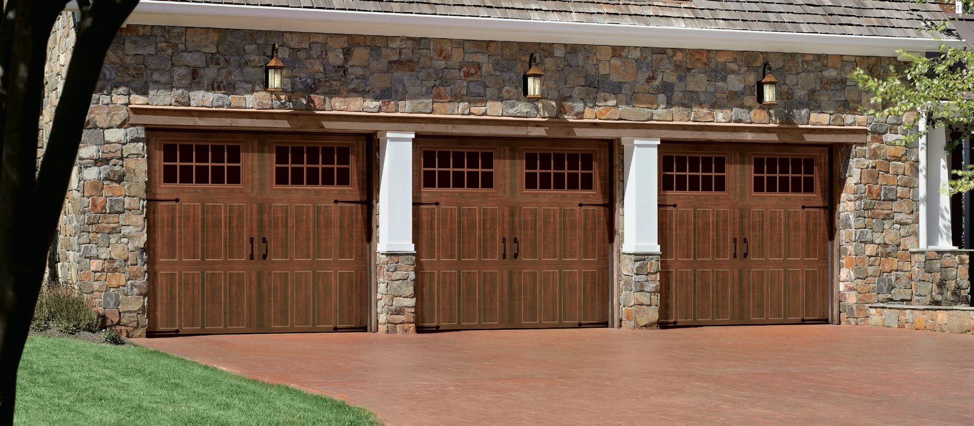 Three brown wooden garage doors on a stone exterior house with a red brick driveway and green lawn