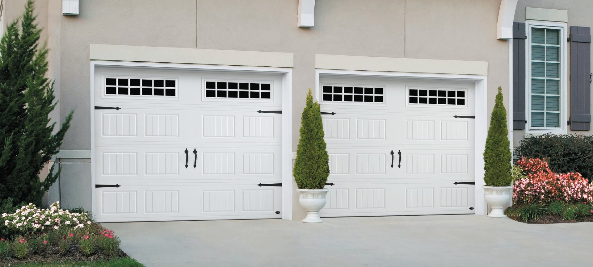 Two white garage doors with black window panes on a beige house facade, flanked by green cypress shrubs and flowering plants
