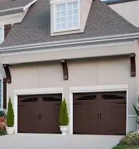 Two dark brown garage doors on a white house with dormant window and landscaping
