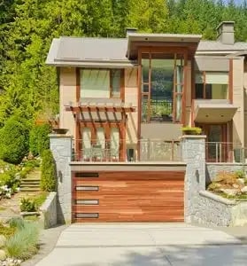 Modern two-story house with wood and stone exterior, horizontal slat garage door, and wooden deck surrounded by green landscaping